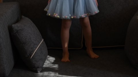 Toddler girl in short blue tutu skirt with barefoot legs jump on couch with pillows. Kid having fun at home. Authentic lifestyle childhood concept