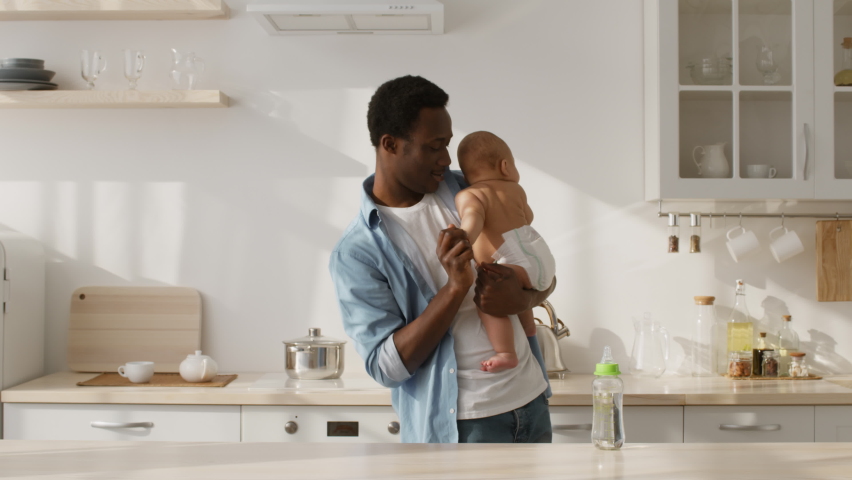 Paternity leave. Happy african american father lullying his newborn baby in diaper, dancing with toddler at kitchen interior, slow motion Royalty-Free Stock Footage #1071521455