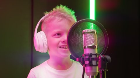 Caucasian kid records new music track, sings song into microphone. Process of recording song in recording studio. Portrait young blond boy singer wearing headphones, singing song in recording studio.