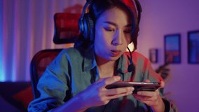 Happy asia girl gamer wear headphone competition play video game online with smartphone colorful neon lights in living room at home. Esport streaming game online, Home quarantine activity concept.