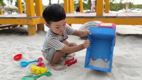 A cute Asian boy playing sand in a sandbox, a little playground, one kid happily playing in the sandpit. Build a sand castle. It's a fun toy. Public park in the summer outdoors