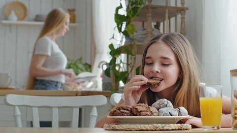 Little girl teenager child daughter schoolgirl sitting at table in home kitchen eating delicious homemade chocolate cookies enjoying breakfast on background of blurred mother washing dishes plate