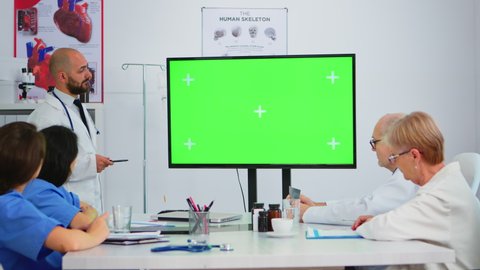 Doctor explaining meeting topics by previewing on a green screen monitor in hospital conference room. Team of doctors with diverse ages brainstorming looking at mock-up display with green chroma key.