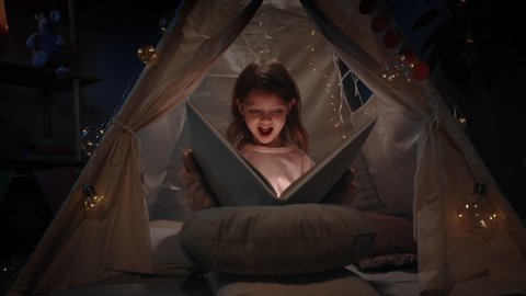 Excited little girl in cute makeshift hut turning pages at home in evening. Cute pretty child sitting on floor while reading book with flashlight. Concept of leisure and careless childhood.