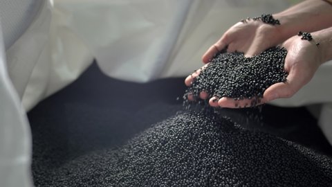 Hands lift black polymer granules from a cloth bag in a garbage recycling plant. Plastic pellets are crumbling or poured from the palms. Raw materials for recycled plastic are used in manufacturing.