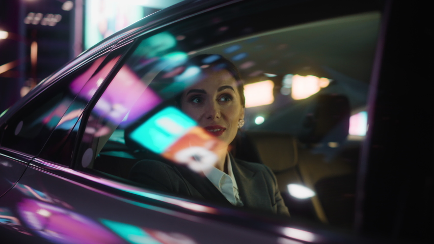 Beautiful Businesswoman is Commuting from Office in a Backseat of Her Luxury Car at Night. Entrepreneur Passenger Traveling in a Transfer Taxi in Urban City Street with Working Neon Signs. Royalty-Free Stock Footage #1071559633