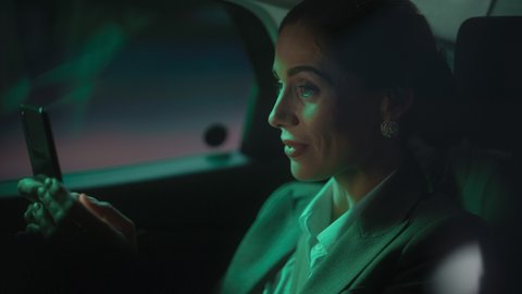 Portrait of Successful Businesswoman Commuting from Office in a Backseat of Her Car at Night. Entrepreneur Using Smartphone while in Transfer Taxi in Urban City Street with Working Neon Signs.