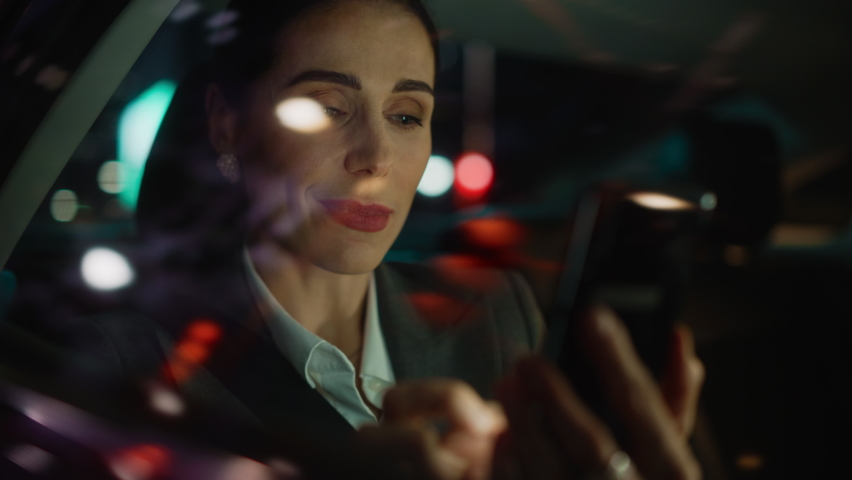 Beautiful Businesswoman is Commuting from Office in a Backseat of Her Car at Night. Entrepreneur Using Smartphone while in Transfer Taxi in Urban City Street with Working Neon Signs.