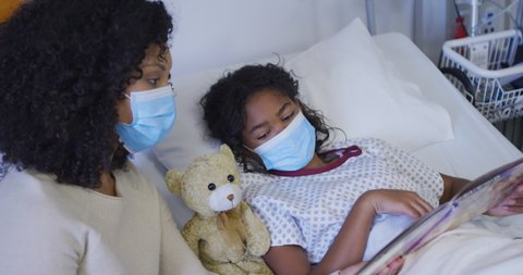 Mixed race mother and daughter lying on hospital bed reading book, both wearing face masks. medicine, health and healthcare services during coronavirus covid 19 pandemic.
