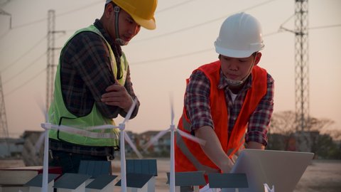 Asian engineer, foreman or leader discussion about home solar cell farm and wind turbine model in construction site project and High voltage power line pylon at sunset. Teamwork, Leadership concept