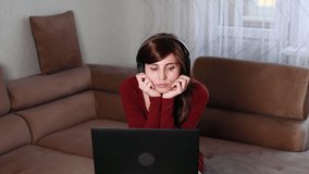 Thoughtful attractive european woman with long dark hair and big brown eyes in headphones in red dress sitting on comfy sofa at home working on laptop computer . Serious student studying at home.