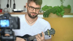 Male smiling blogger shoots video on camera, playing guitar at home. Online music teaching concept. Ukulele