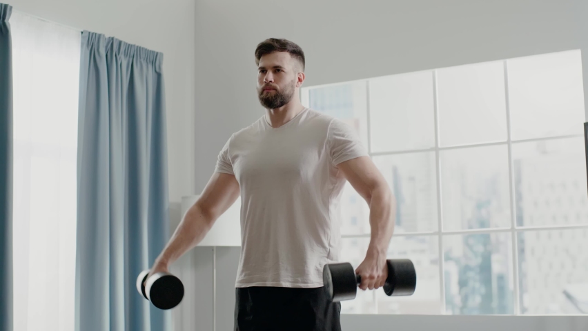 Concentrated muscular bodybuilder man with beard practices lateral lift holding black dumbbells standing at home slow motion Royalty-Free Stock Footage #1071575812