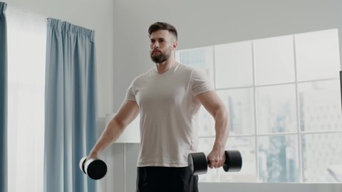 Concentrated muscular bodybuilder man with beard practices lateral lift holding black dumbbells standing at home slow motion