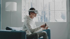 Confused man in white hoodie and virtual reality glasses moves hand with controller and smiles on couch by window slow motion