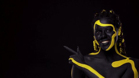 Face art. Girl pointing on copy space with fingers. Woman with black and yellow body paint. Young african girl with colorful bodypaint. An amazing afro american model with makeup.