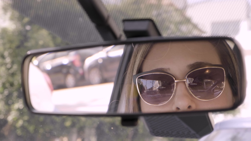 Woman reflected in car rearview mirror. closeup of young beautiful lady looking in mirror taking down sunglasses and smiling to passenger. Driver flirting with guy in the back seat of car. Traffic jam