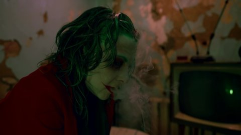 cosplay on clown, actor with makeup and suit is smoking in old flat, exhaling smoke and grimacing