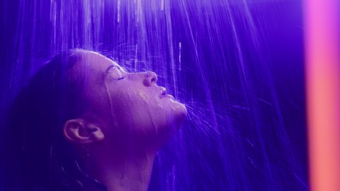 Young woman takes a shower in colourful filter of vibrant ultra-violet light. Water fall in multi-color neon lighting of dark bath room. Calm face of pretty lady or confident girl in bathroom closeup