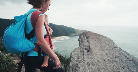 Family hiking. Mother hikes with baby in the mountains and achieves the top. Young woman hiker carries her baby in wrap sling and walks in the mountains