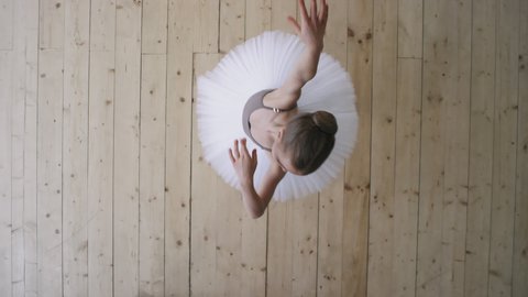 Top-view shot of pretty little ballerina in white tutu dress dancing in empty ballet studio whirling and twirling performing pirouettes