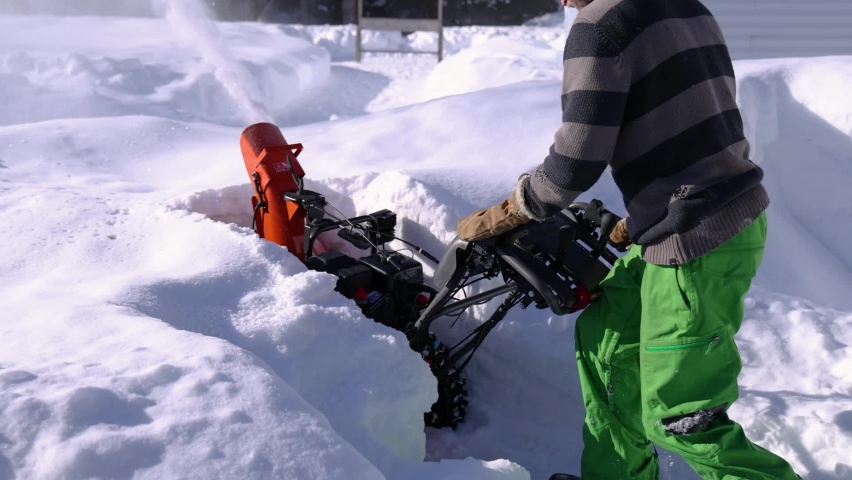 Close up shooting of heaps of fresh snow being removed and shot in the air by a bright orange mechanical snowplough operated by a young worker.
