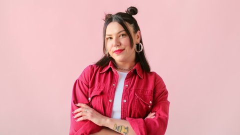 Stylish brunette woman with tattoo blinking eye, smiling and looking at camera on pink background. Winking and flirting trendy female.