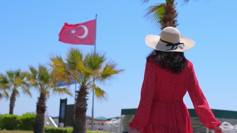 Traveler woman traveling in red dress, hat walking with suitcase at luxury resort on background flag of Turkey blue sky. Female tourist woman in hotel. travel tourism beach vacation happy holiday