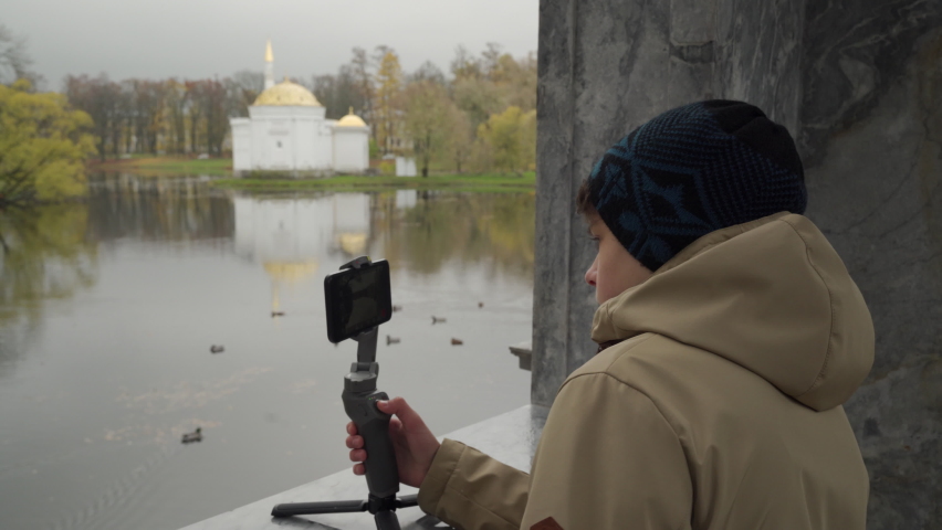 Vlogger boy with mobile phone camera stabilizer recording outdoor video blog in autumn park. Teenager shooting video for travel vlogging. Catherine Park, Tsarskoye Selo, Saint Petersburg, Russia.