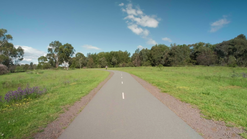 Young man riding a skateboard going very fast around a corner along a road in a park with green grass, trees and cloudy blue sky in the background.