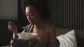 A black happy woman having breakfast cereal with milk and holding a plate while sitting on the bed in the room - Powered by Shutterstock - Get 15% off with code: PIKWIZARD15