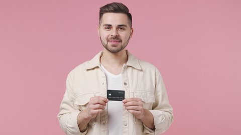 Handsome happy young man 20s wears light jacket white t-shirt pointing index finger on mockup plastic credit bank card showing thumbs up isolated on pastel pink wall color background studio portrait