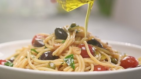 Chef pours olive oil over spaghetti alla puttanesca, served on a plate.