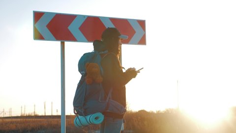 A young woman traveler uses her phone to choose the right road. Hiker woman with backpack choose in a road fork between two different path directions at the sunset. Concept of choose the correct way.
