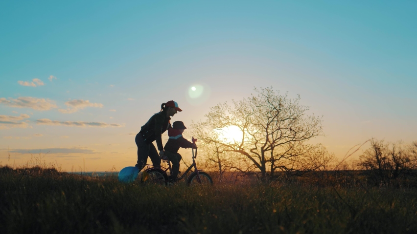 Childhood bike concept. Mother teaching son to ride bicycle. Happy cute boy learn to riding a bike in park at sunset time. Young mom teaching son to ride bike first time on countryside rural road. Royalty-Free Stock Footage #1071645259