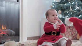 authentic cute chubby infant baby girl in santa hat and red dress near fireplace with young woman mom celebrating happy new year near christmas tree. Childhood, Parenthood, hollyday, winter concept.