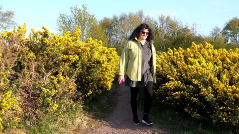 Smiling woman with glasses and long black curly hair walking towards camera POV between gorse bush flowers.