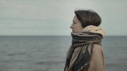 Side view portrait of woman with tousled hair in warm coat and scarf standing against ocean. Close up portrait of young woman with dark hair blowing in wind looking away over ocean