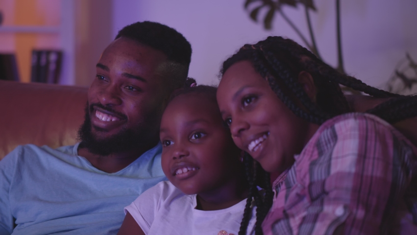 Close up of happy african mother, father and daughter watching tv on couch at home. Positive afro-american young parents and kid relaxing together in evening and enjoying tv series | Shutterstock HD Video #1071652348