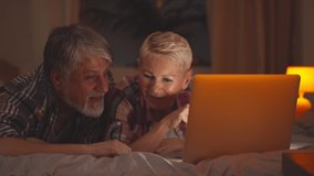 Happy senior couple relaxing on bed and shopping online on laptop in evening. Portrait of cheerful aged husband and wife surfing internet or watching movie on computer in bedroom - Powered by Shutterstock - Get 15% off with code: PIKWIZARD15