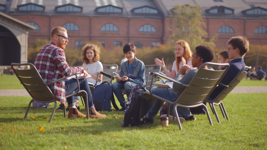 Curious diverse kids sitting with workbooks on chairs and applauding while listening to teacher at outdoor lesson. Young educator and schoolkids siting in circle on schoolyard lawn