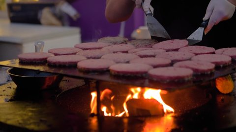 Super slow motion: chef grilling fresh meat cutlets for burgers on brazier with hot flame at summer local food market - close up view. Outdoor cooking, gastronomy, cookery, street food concept
