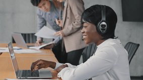 Smiling african female call center agent wear wireless headset using laptop looking at screen consulting customer with online problem make video call in office, black woman operator support service