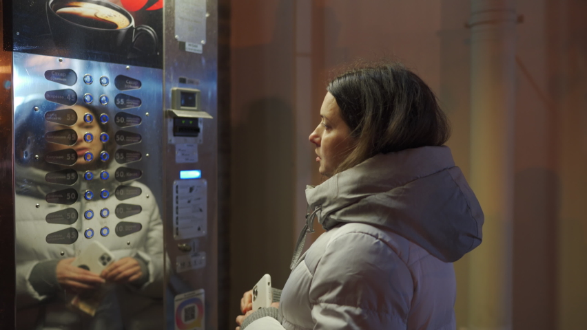Woman buying coffee from vending machine and pushing some buttons on an empty street at cold night. Pavlovsk Park and Palace in autumn, St. Petersburg, Russia.