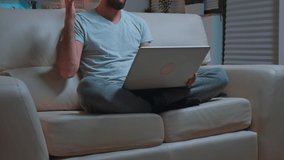 Focused man sitting on sofa in pajamas while talking with collegue during videocall conference using laptop computer. Caucasian male working at communication project late at night in kitchen