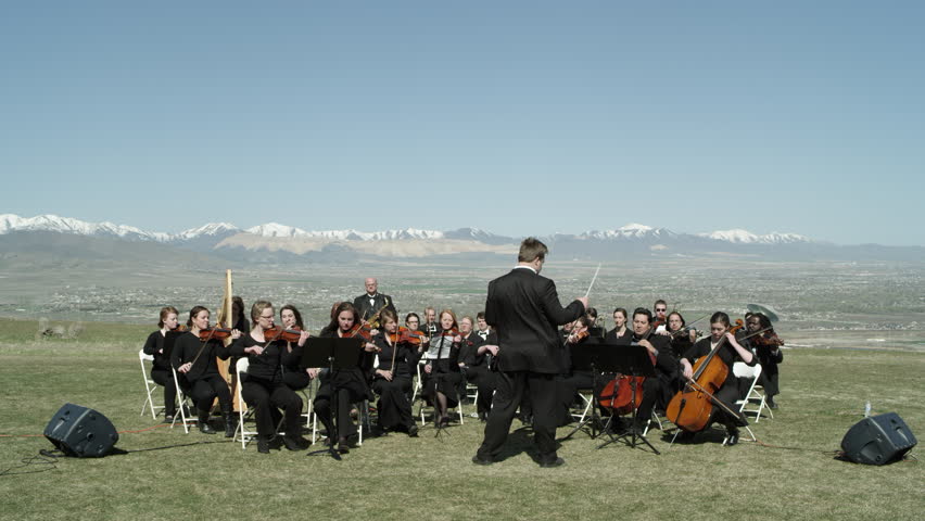 Orchestra performing outdoors on a hill overlooking a valley.