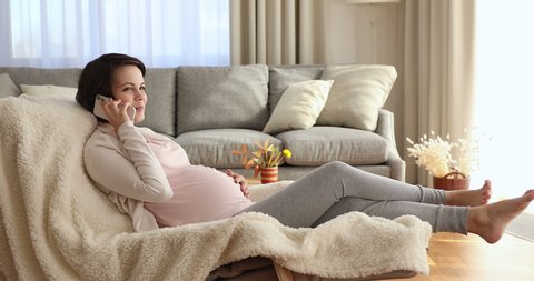 Happy carefree pregnant woman relax on soft cozy armchair in modern living room enjoy pleasant conversation on phone. Share news with family remotely, chat with caring husband while rest alone at home