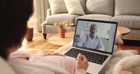 Older male doctor provide online consultation to woman laptop screen view over pregnant female shoulder, relaxing alone at home receiving obstetrician gynaecologist help via video conferencing apps