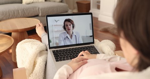 Pregnant woman relaxing at home use laptop receive online consultation from female gynaecologist through video conference, computer screen view over patient shoulder. Medical care by videocall concept