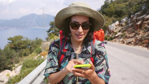 Portrait of Young Hiker Woman Using Mobile Phone Navigation or Social Media App. Girl with a Backpack Walks Down the Road and Looks at her Phone. 4K Slowmotion
